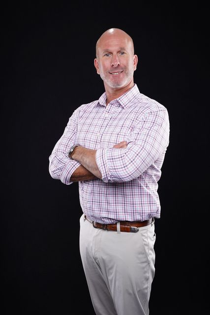 Man with shaved head, crossed arms, wearing a patterned button-down shirt and khakis, standing against a black background.
