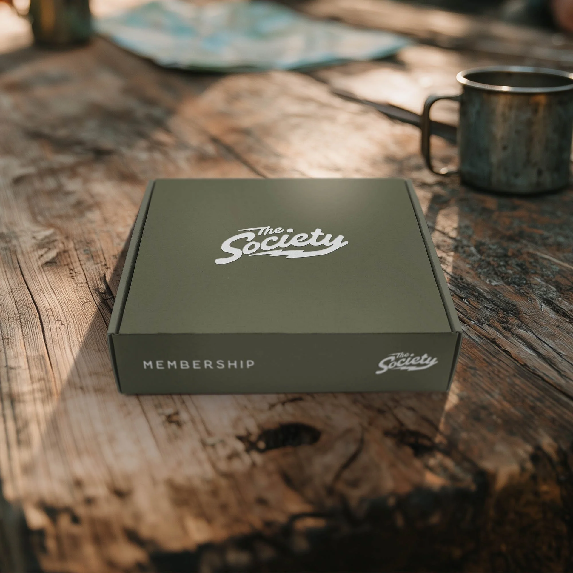 Green "The Society" membership box on a rustic wooden table beside a metal mug