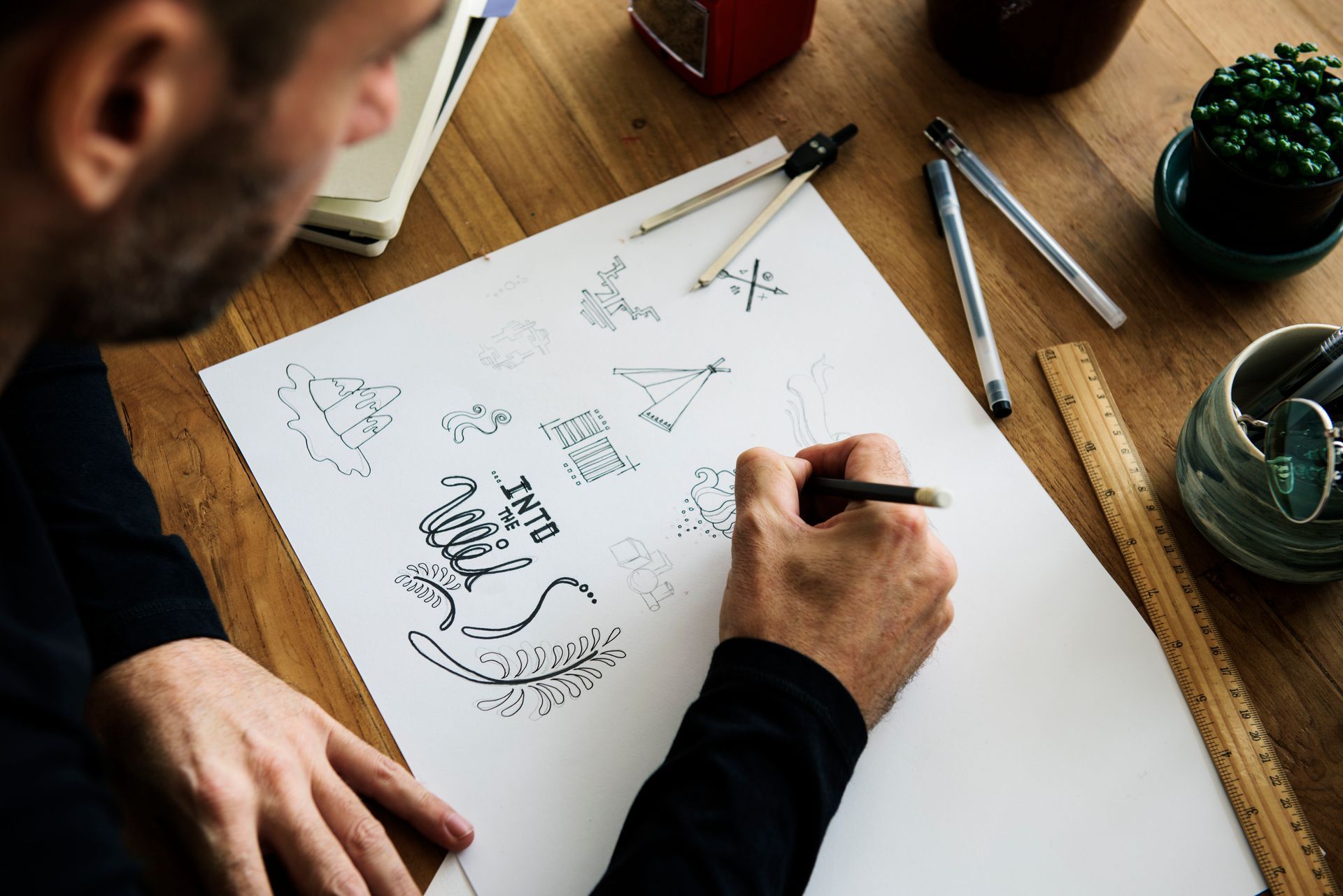 Person sketching design ideas on a white sheet at a wooden desk with pens and a coffee cup.