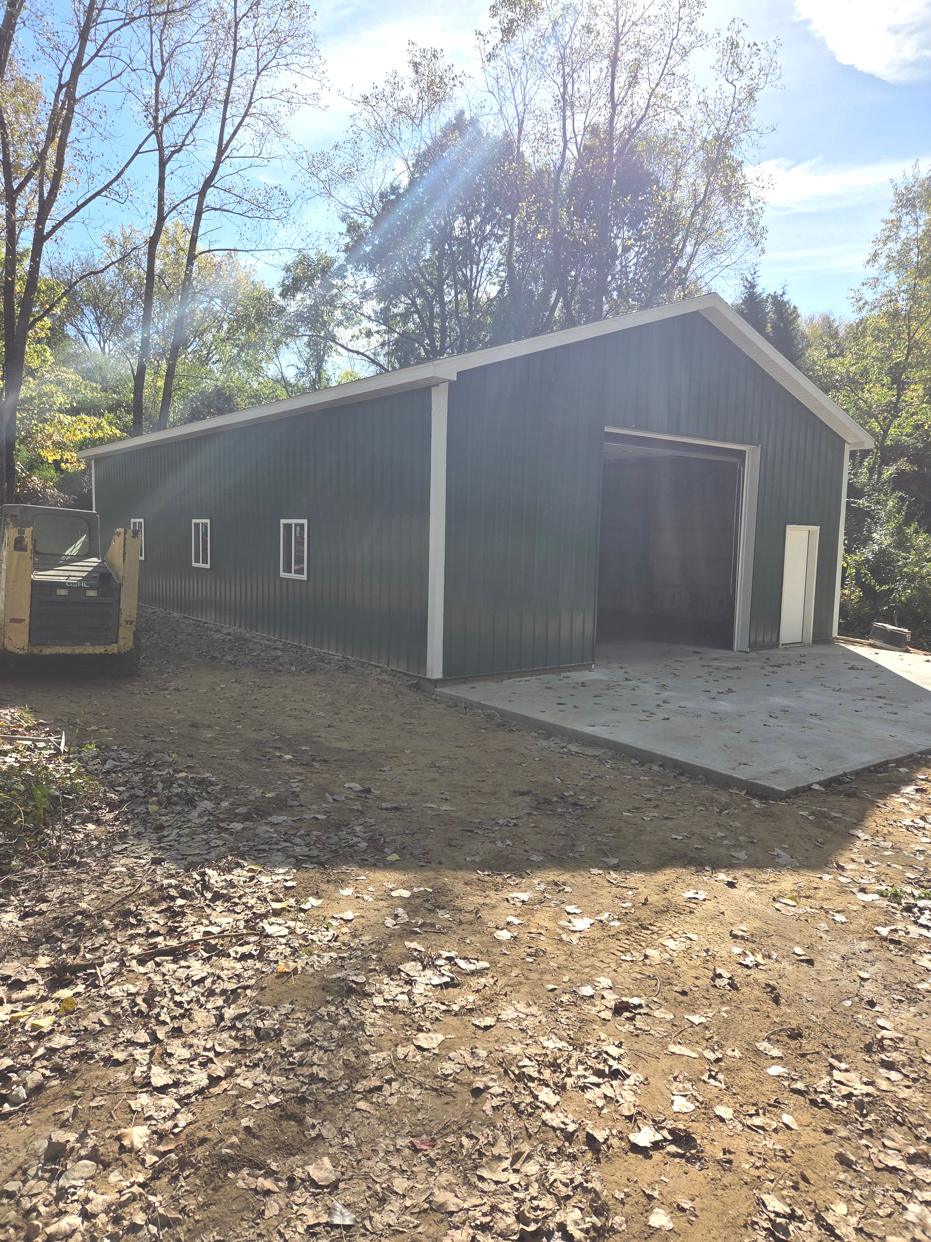 Green metal barn with open doorway and concrete pad, set in a wooded area.