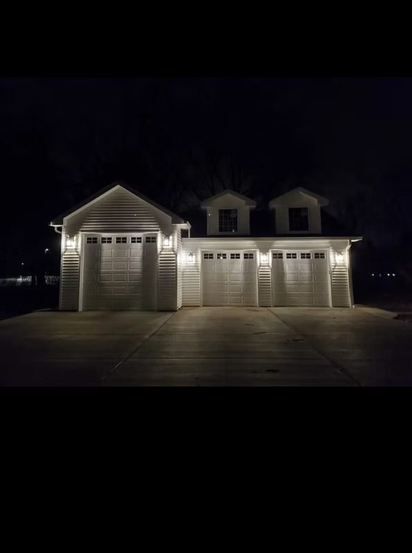White double garage doors with black hardware. Grey driveway, brown building exterior, and a few plants visible.