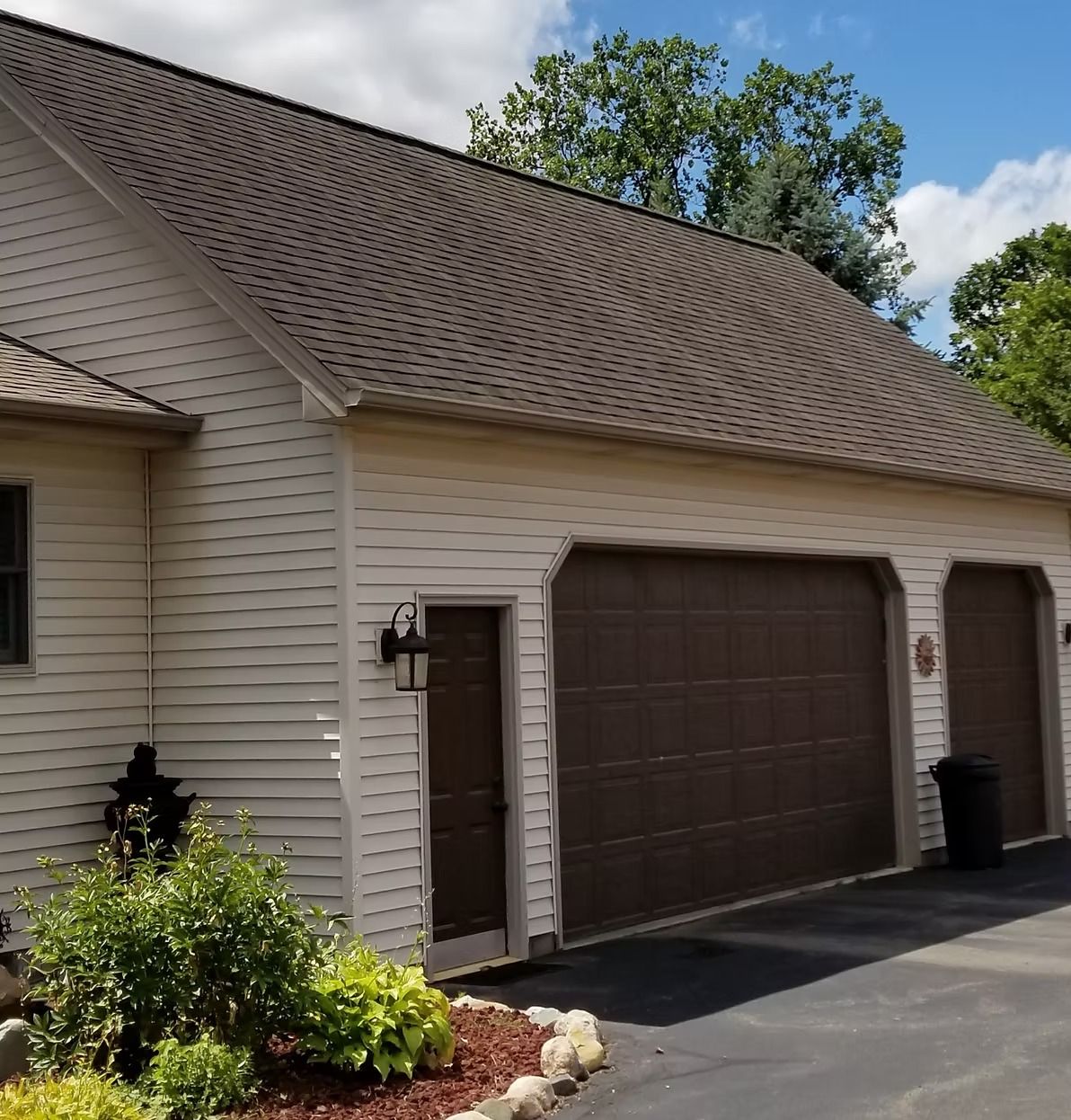 Tan-sided building with brown roof and garage doors; green bushes in front, blue sky in background.
