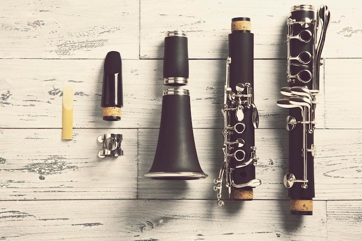 Disassembled clarinet on a light wood surface, parts arranged: reed, mouthpiece, barrel, body, bell.