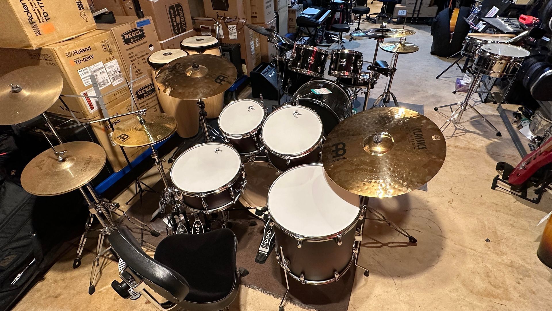 Drum set in a cluttered room. Multiple drums, cymbals, and boxes. Brown and black colors dominate.