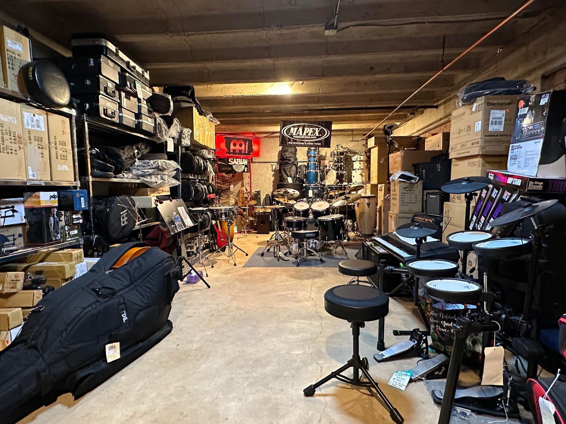 A cluttered music equipment storage room. Drums, keyboards, boxes, and a cello case fill the space.