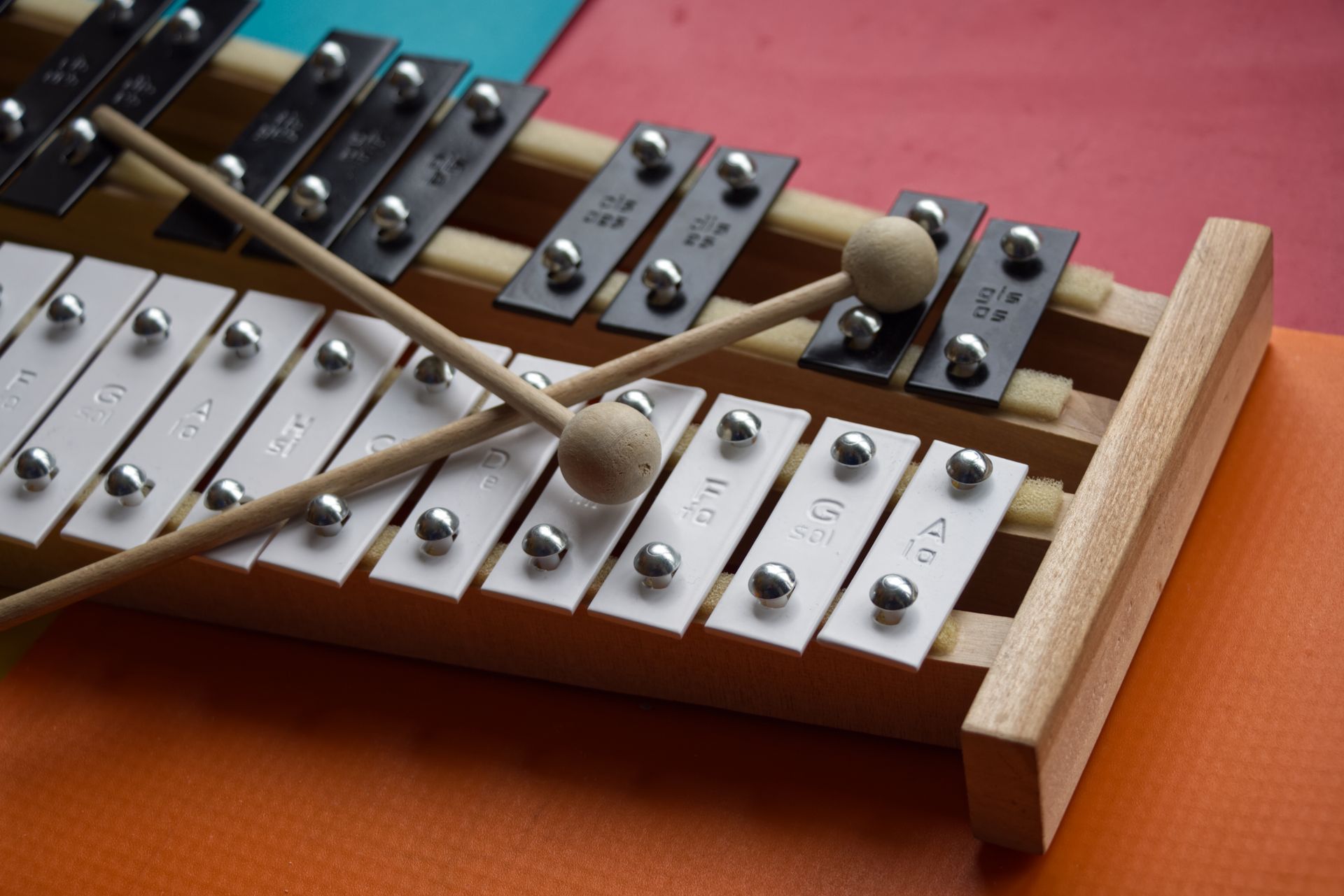 Xylophone with mallets, white and black metal bars, wood frame, resting on orange and blue surfaces.