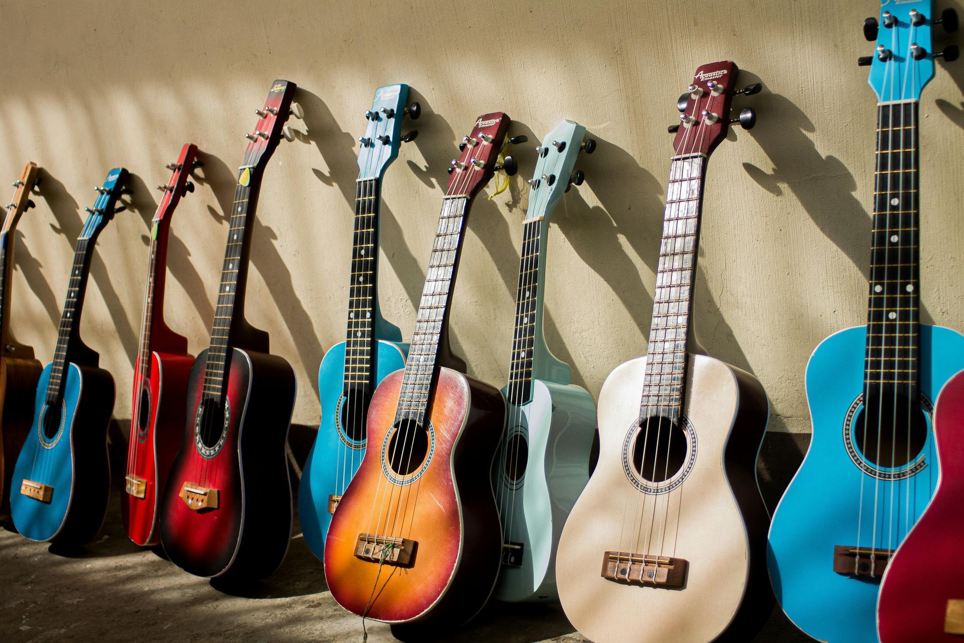 Row of colorful ukuleles leaning against a beige wall, casting shadows.