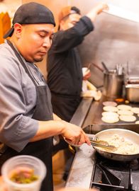 A man is cooking food in a pan on a stove in a kitchen.