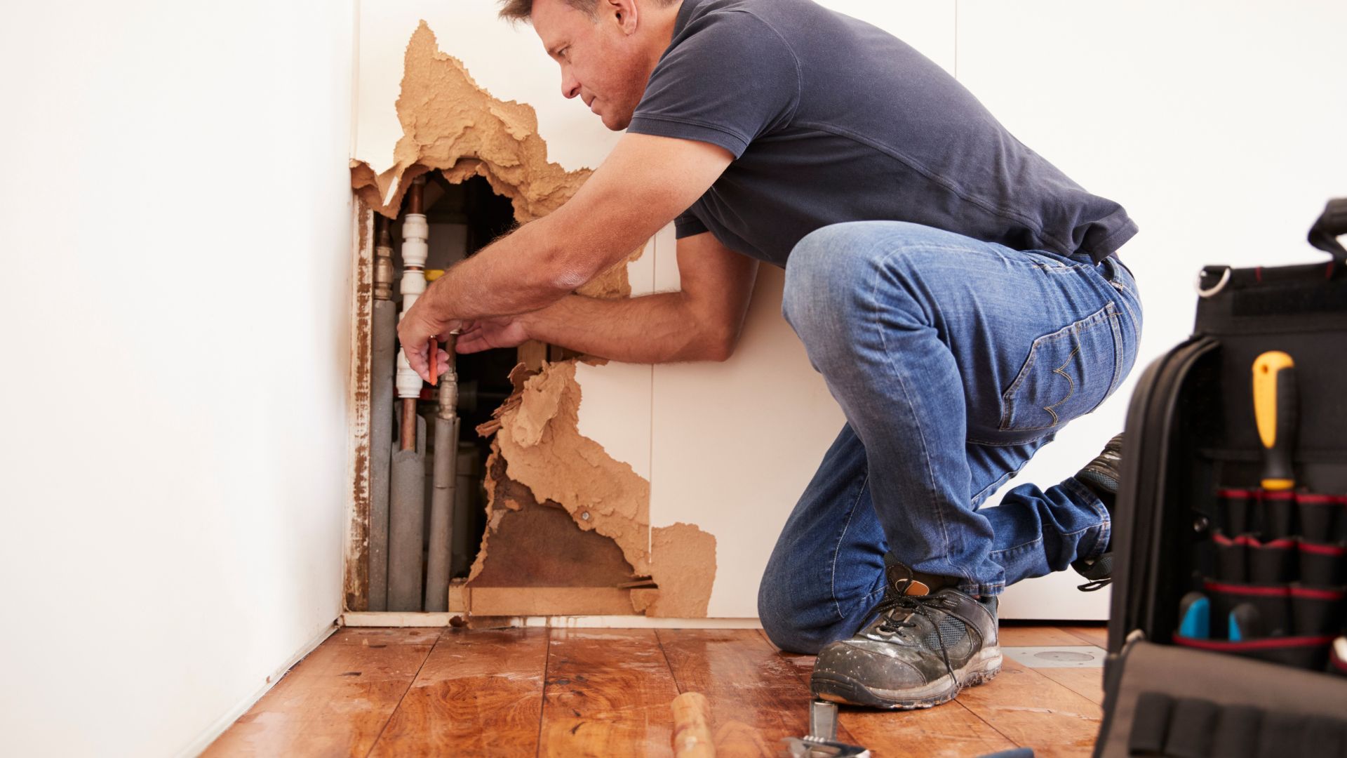 Plumber kneels by damaged wall, repairing pipes with tools and water on the floor.