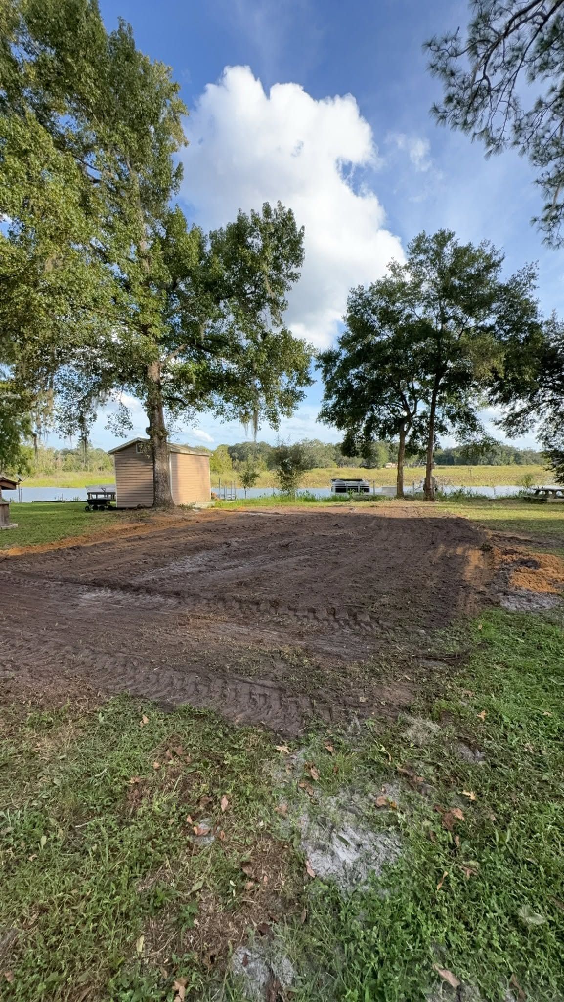 Grassy park with trees, a dirt clearing, and a lake in the background under a partly cloudy sky