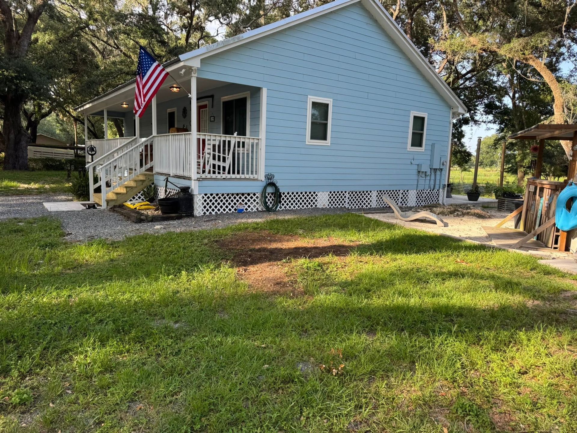 Light blue cottage with white porch and an American flag, set in a grassy yard with trees.