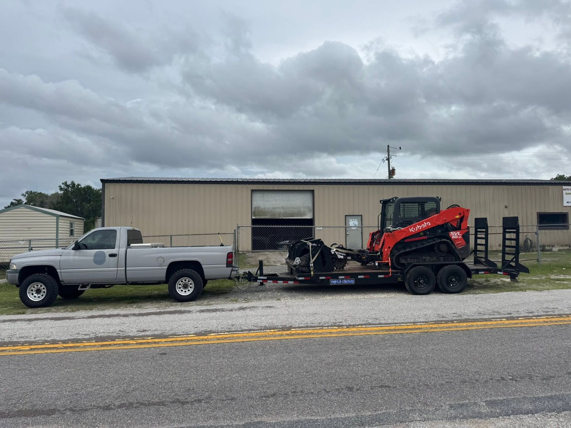 Pickup truck towing red skid steer on trailer outside a beige industrial building under cloudy sky