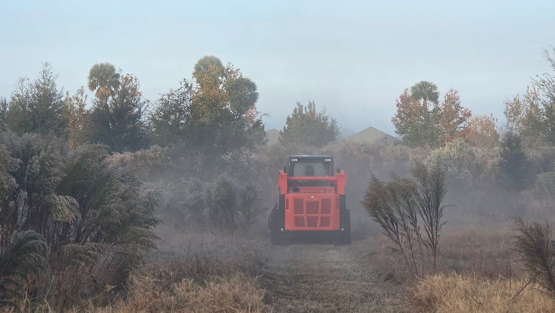 Red tractor driving through a dusty field with shrubs and trees in the background