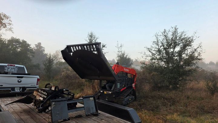 An orange excavator crushed in a ditch beside a pickup truck, with debris and trees around it.