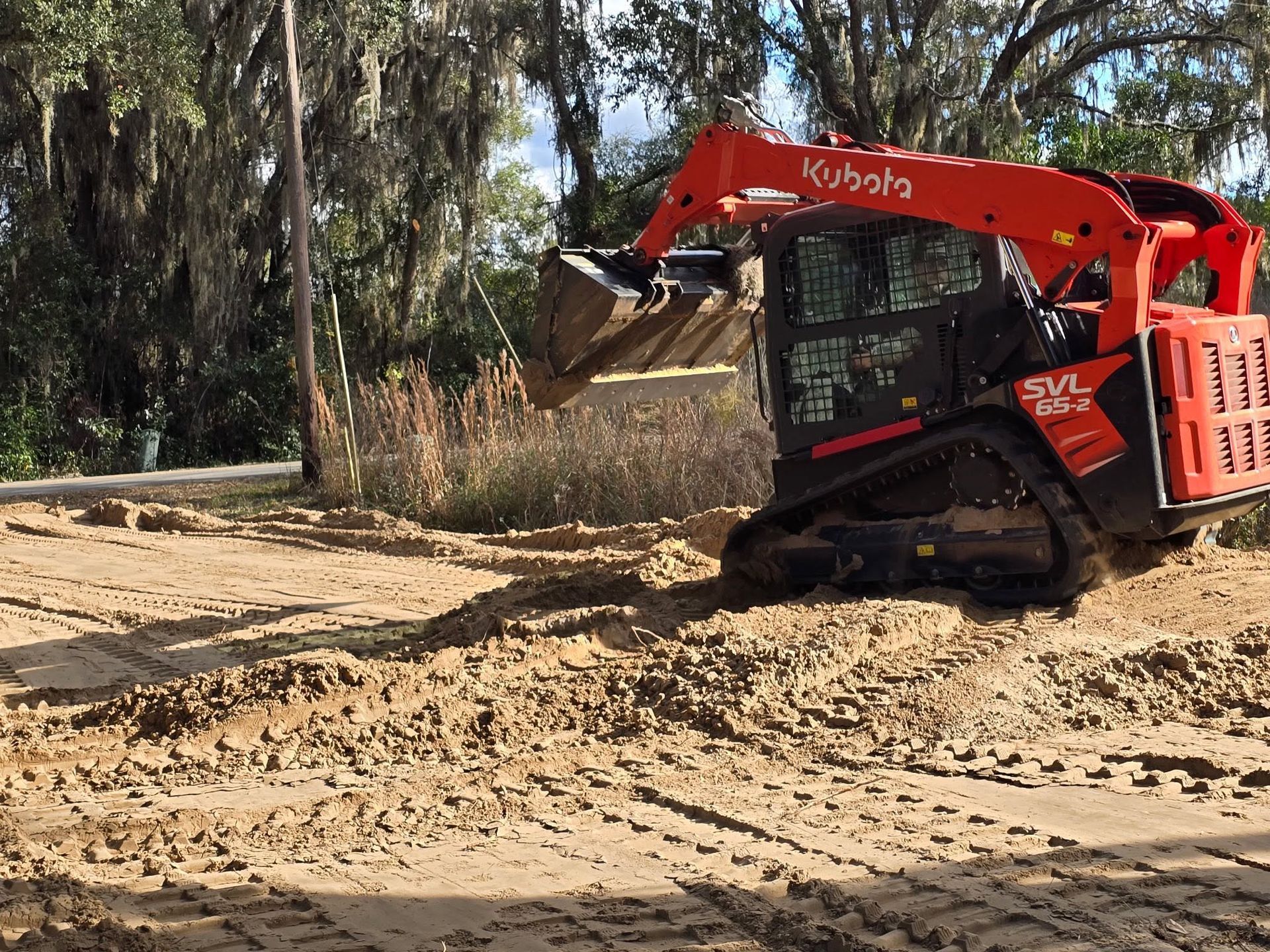 Red Kubota skid steer leveling dirt on a muddy construction site near trees