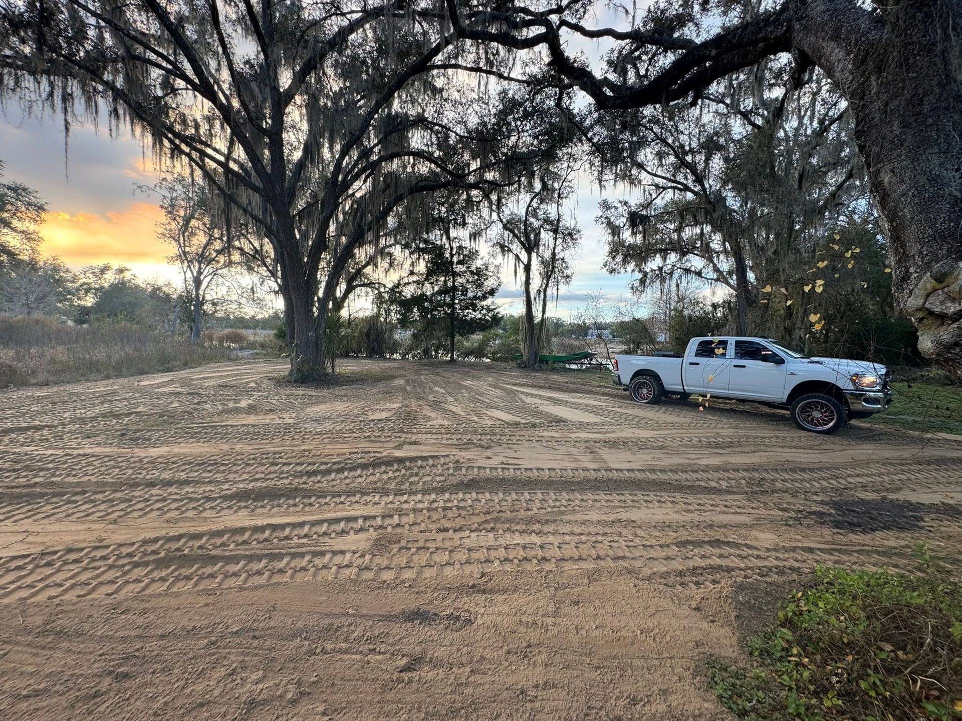 White pickup truck parked on a sandy dirt clearing beneath large trees at sunset