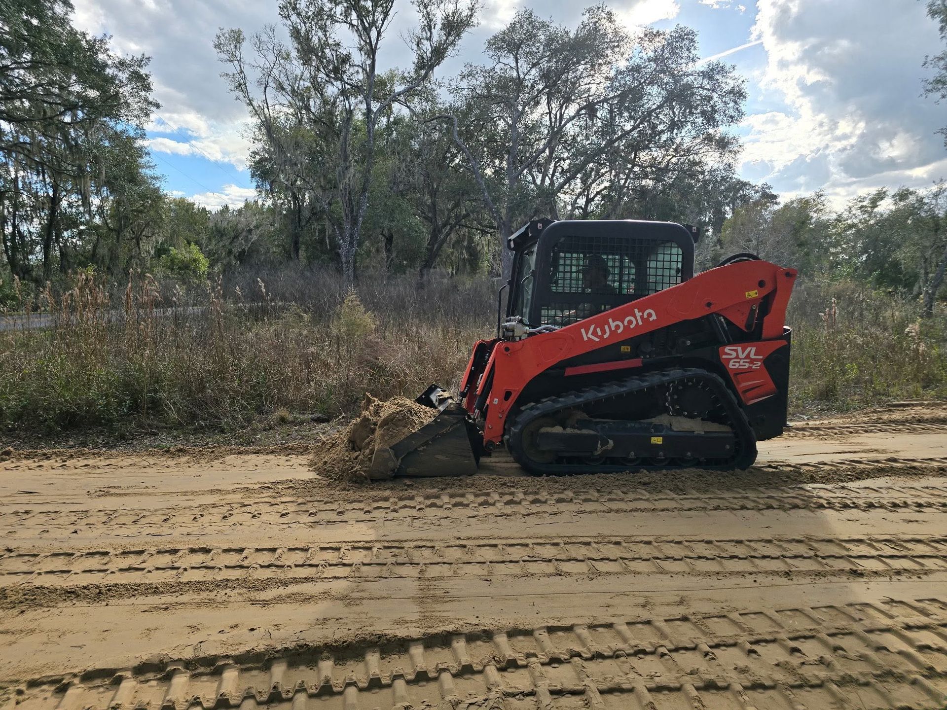 Red skid-steer loader pushing dirt on a sandy road in a wooded area.