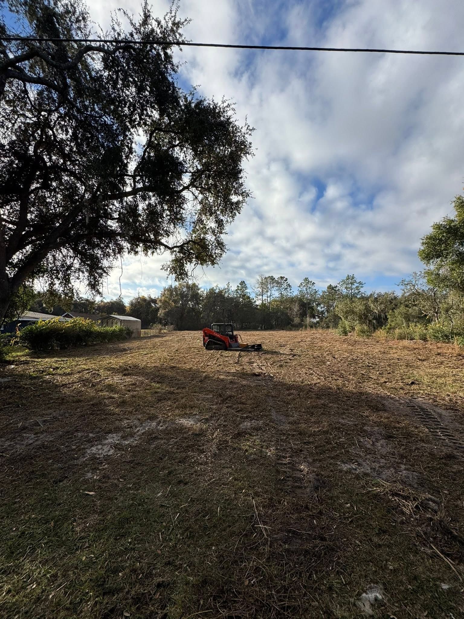 A red lawn mower sits in a sunlit field beneath a cloudy sky, with trees framing the scene.