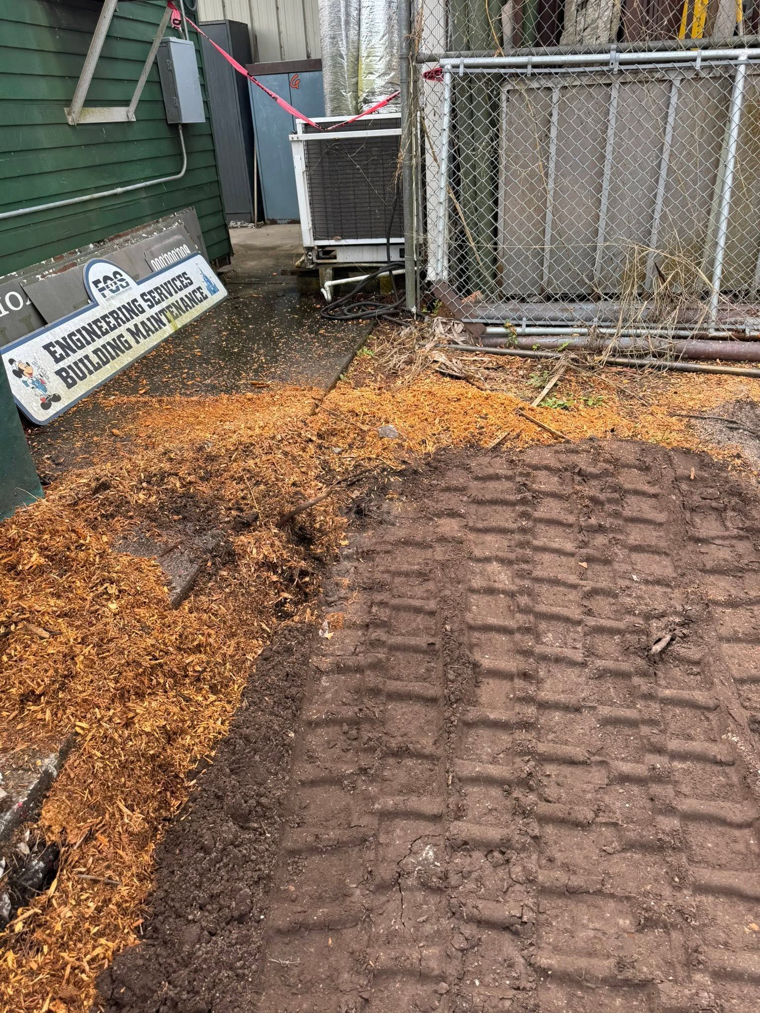 Muddy gravel path beside a green building and chain-link fence, with a brick walkway in the foreground.