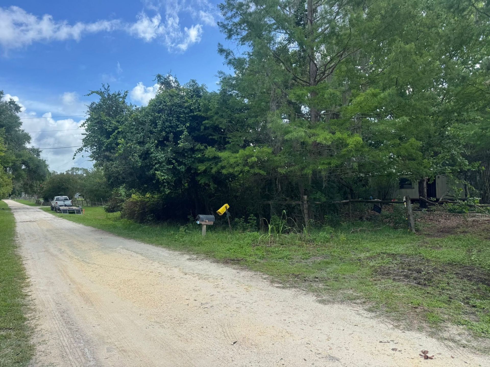 Dirt road beside green trees and a fenced yard under a partly cloudy blue sky