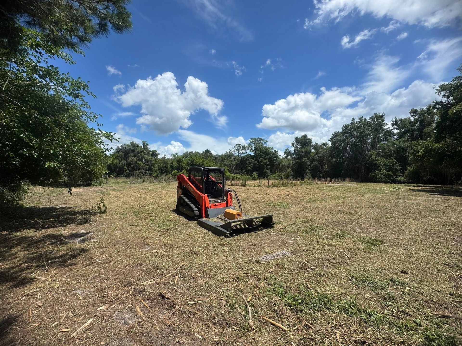 Orange skid steer clearing a field under a blue sky with clouds