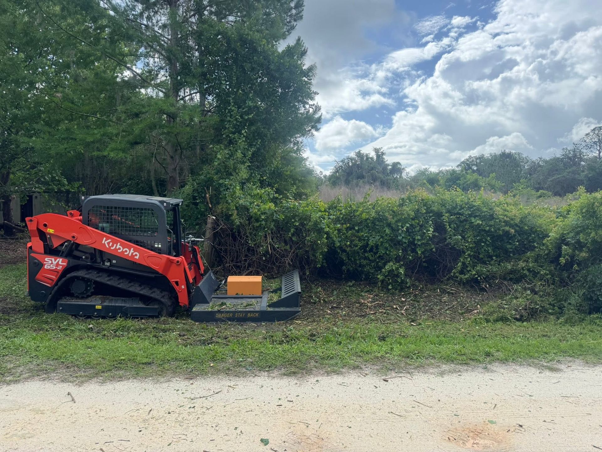 Red skid steer near hedges on a dirt path under cloudy sky