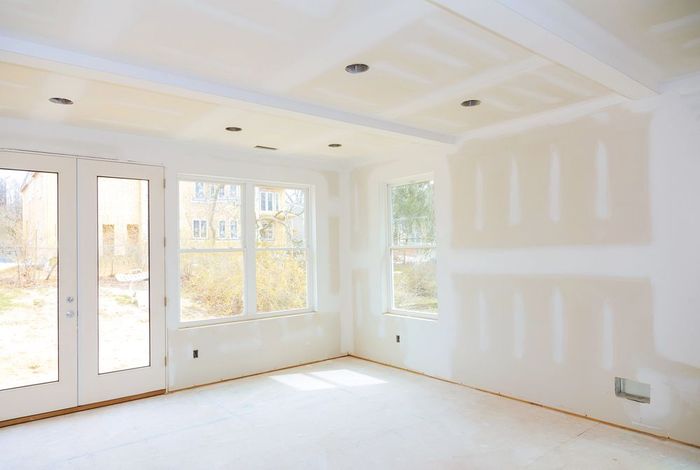 Stanza luminosa e vuota, con pareti bianche, luci a soffitto, ampie finestre e riparazioni non finite nel cartongesso