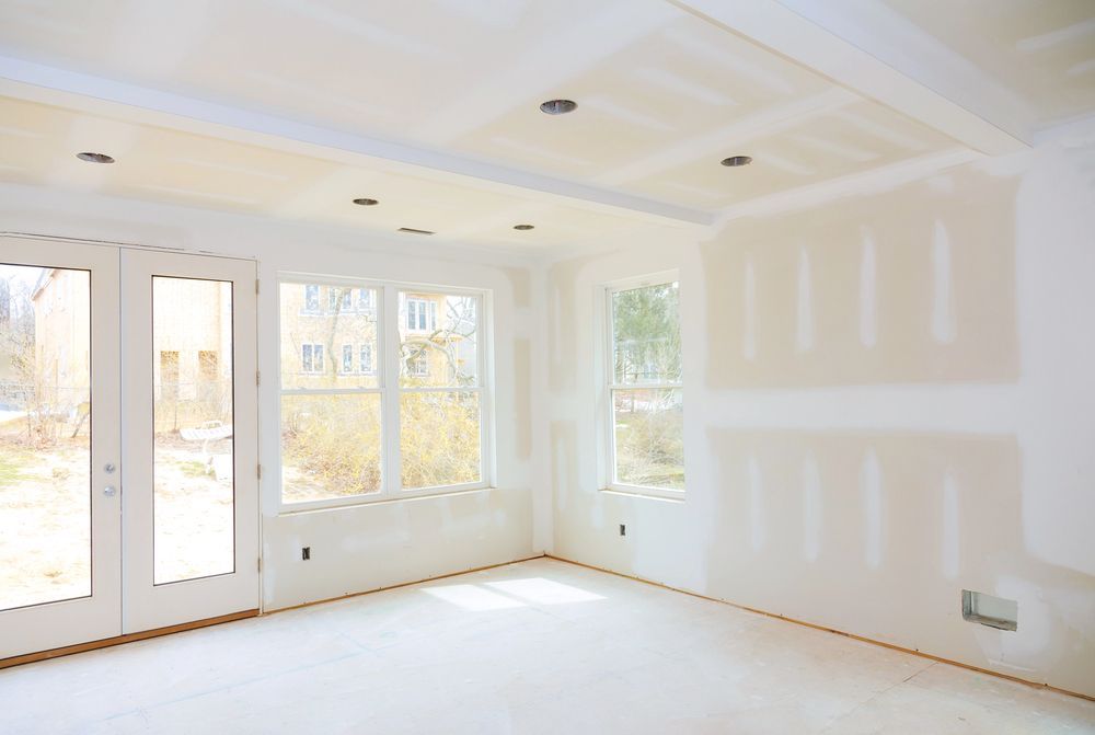 Stanza luminosa e vuota, con pareti bianche, luci a soffitto, ampie finestre e riparazioni non finite nel cartongesso