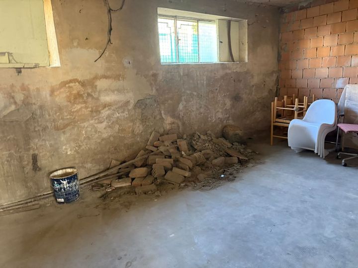 Stanza non finita con parete di mattoni a vista, cumulo di detriti, finestrella e secchio sul pavimento di cemento.