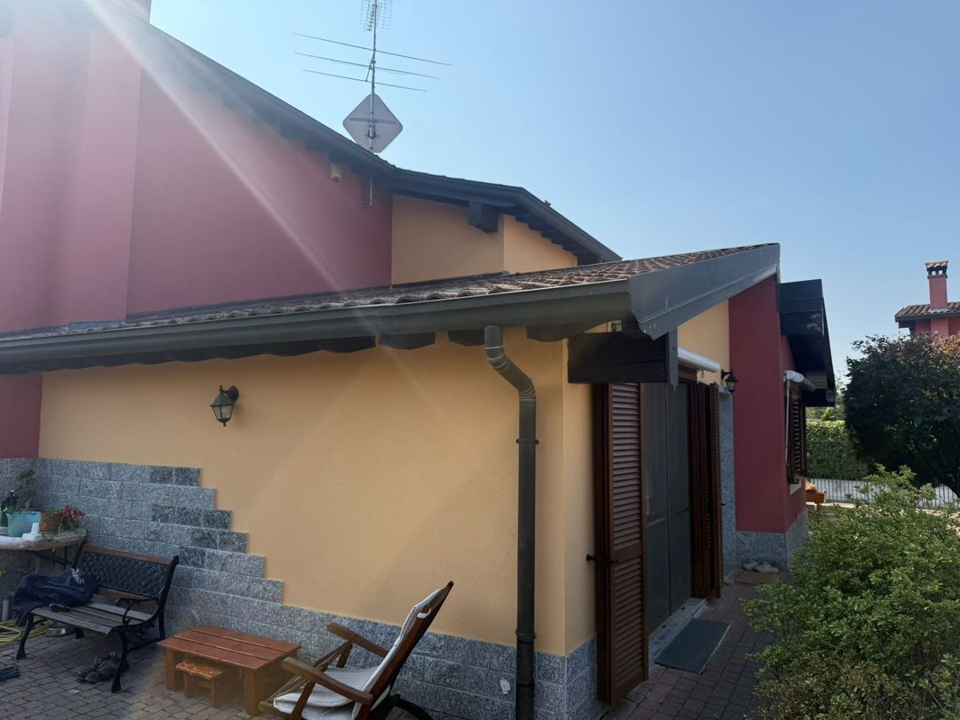 Piccola casa gialla con il tetto e la tenda da sole rossi, gradini in pietra e posti a sedere all’aperto in un giardino soleggiato