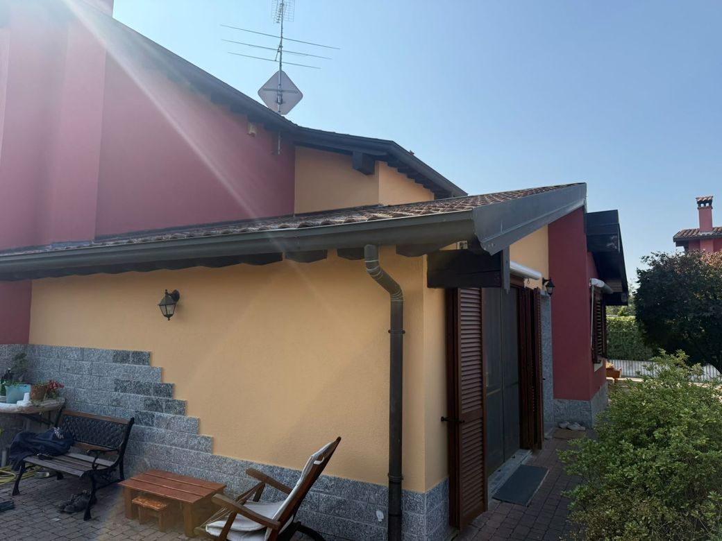 Esterno di una casetta con pareti beige, tetto rosso, gradini in pietra e un patio coperto con sedie