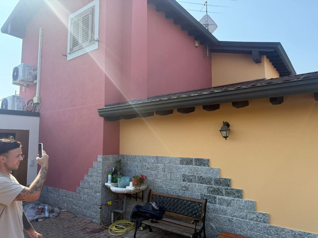 Persona che fotografa l’esterno di una casa rosa e gialla, con una panchina e un muro di pietra in un cortile