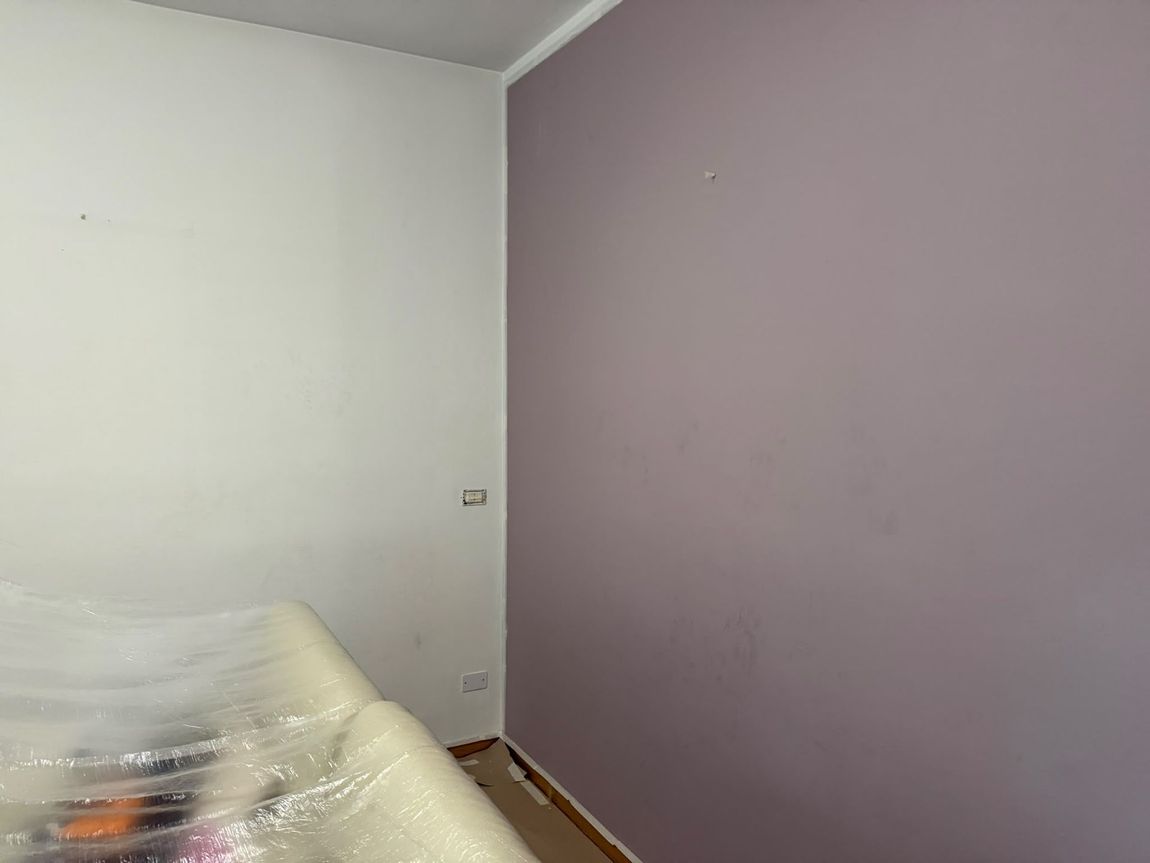 Stanza vuota con pareti bianche e viola chiaro, un letto avvolto nella plastica in un angolo e il pavimento spoglio.