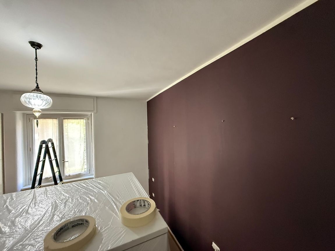 Stanza con una parete d’accento color bordeaux scuro in fase di tinteggiatura, con un telo protettivo e una scala vicino alla finestra.