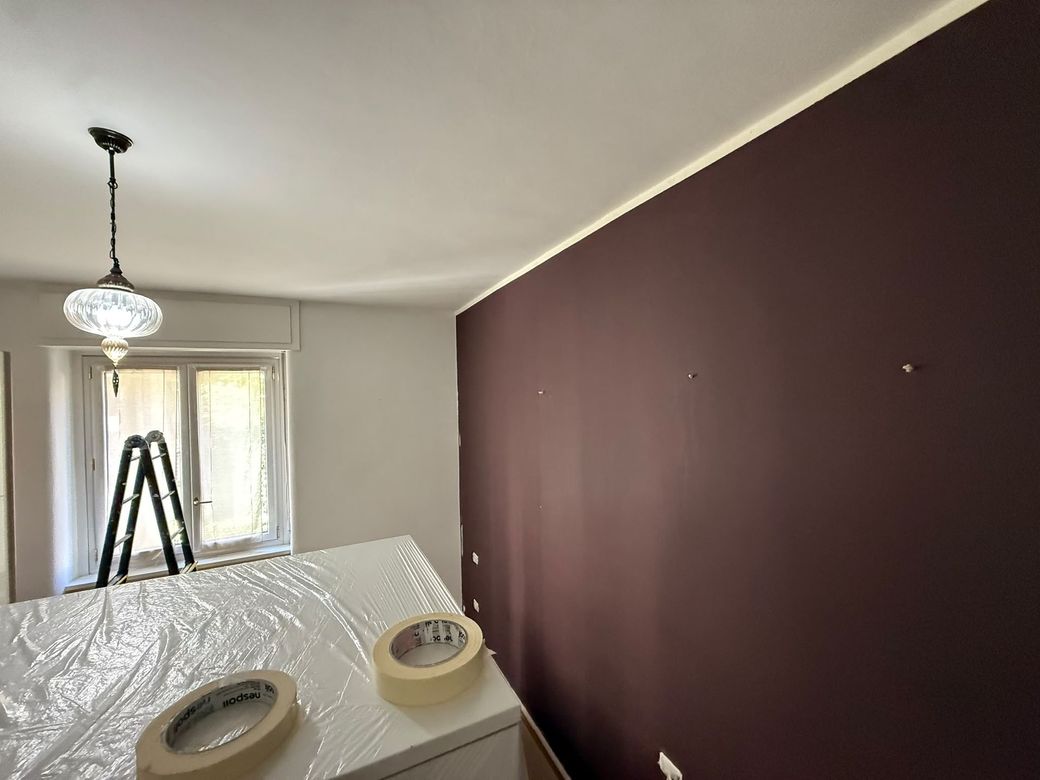 Stanza con una parete d’accento color bordeaux scuro in fase di tinteggiatura, con un telo protettivo e una scala vicino alla finestra.