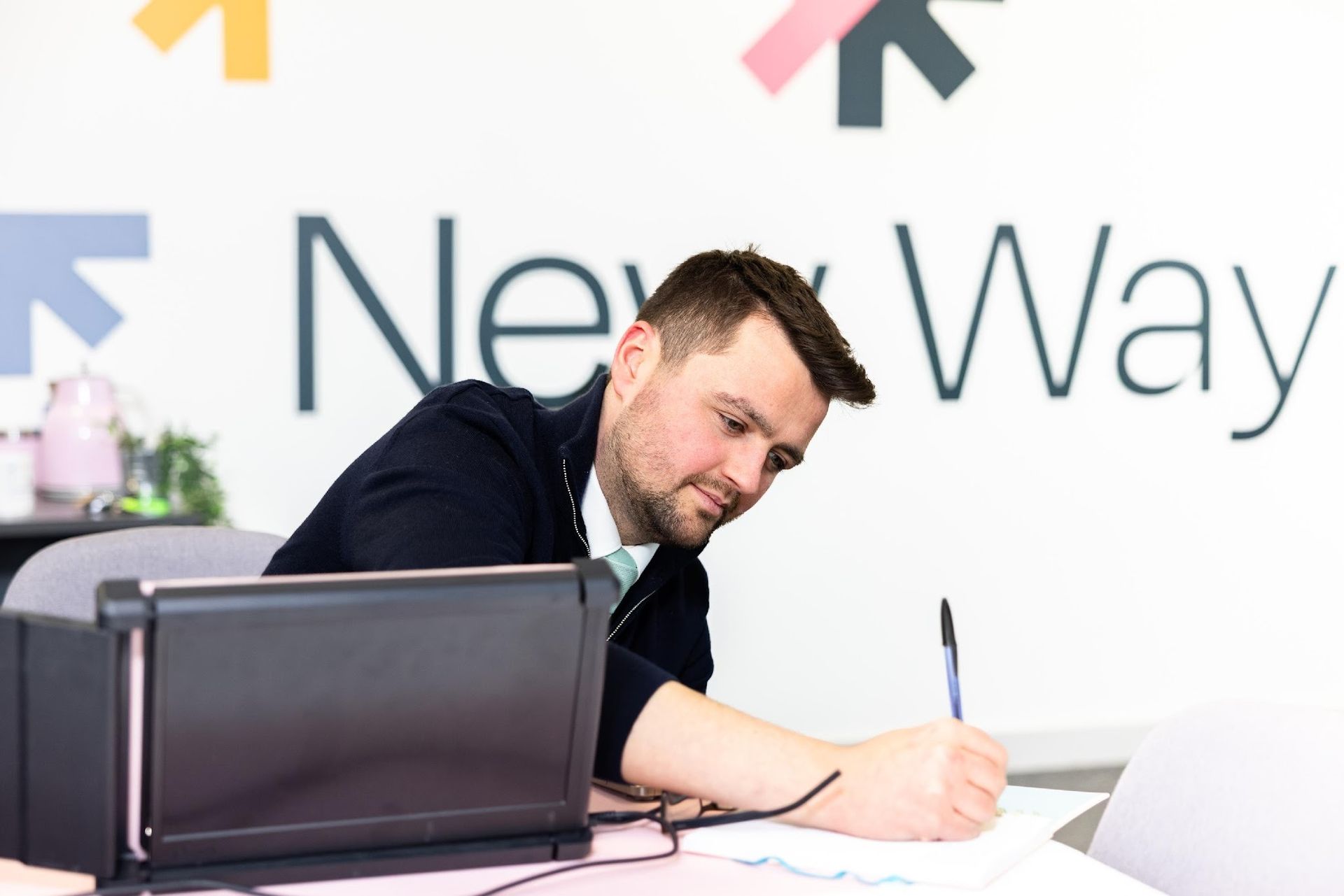 Man signing paperwork at a desk beside a laptop in a bright office, with “New Way” on the wall