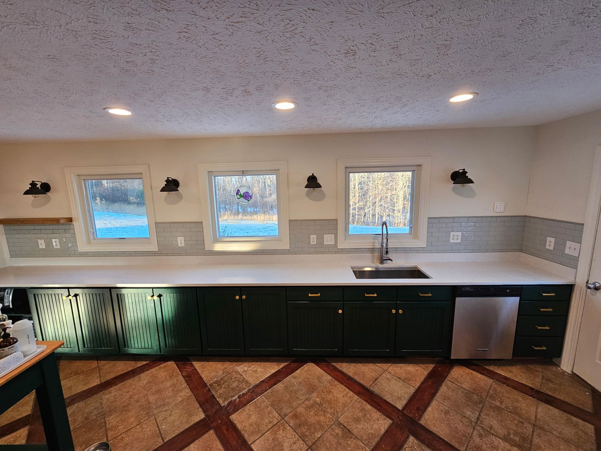 A kitchen with dark green cabinets, white countertops, three windows, a stainless dishwasher, and brown patterned tile floor.