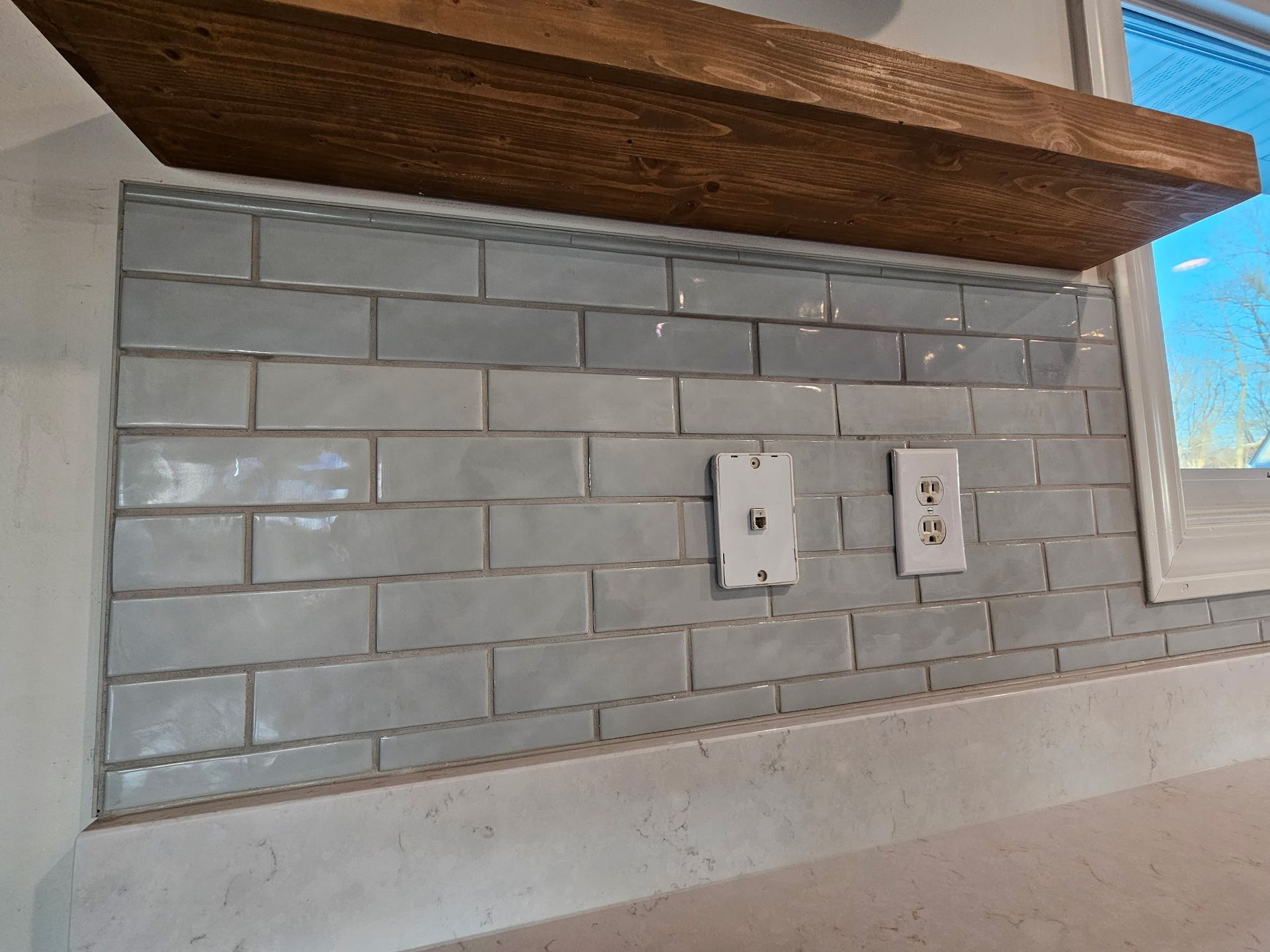 Light gray subway tile backsplash under a rustic wooden shelf, featuring two wall-mounted electrical outlets.