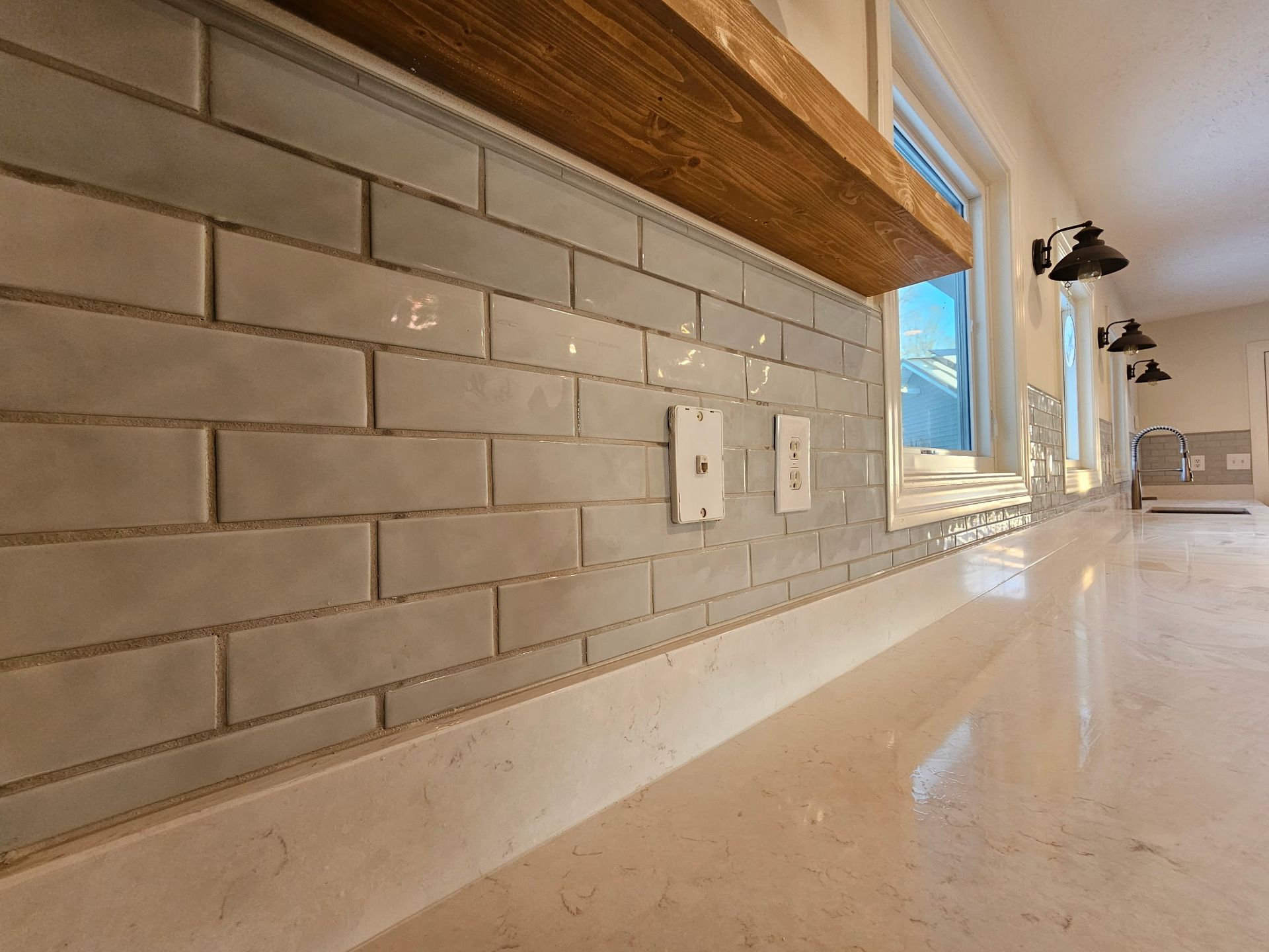 A kitchen backsplash with light grey subway tiles, a wooden floating shelf, and a light-colored countertop.