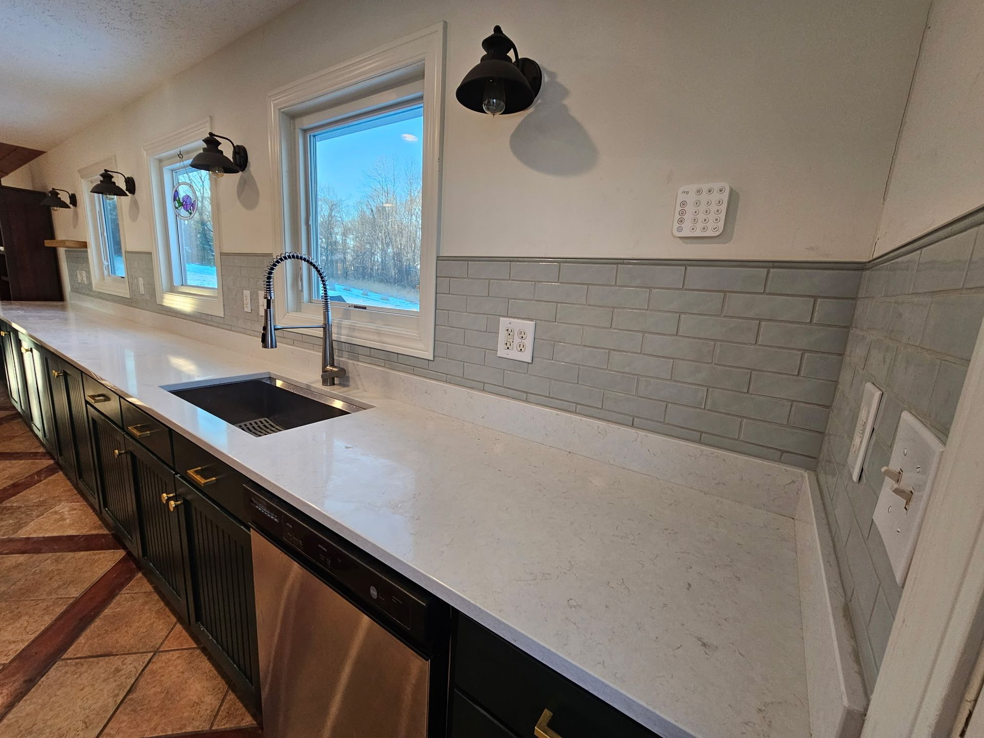 A kitchen counter with a white quartz countertop, black cabinets, and a grey brick-style backsplash under two windows.