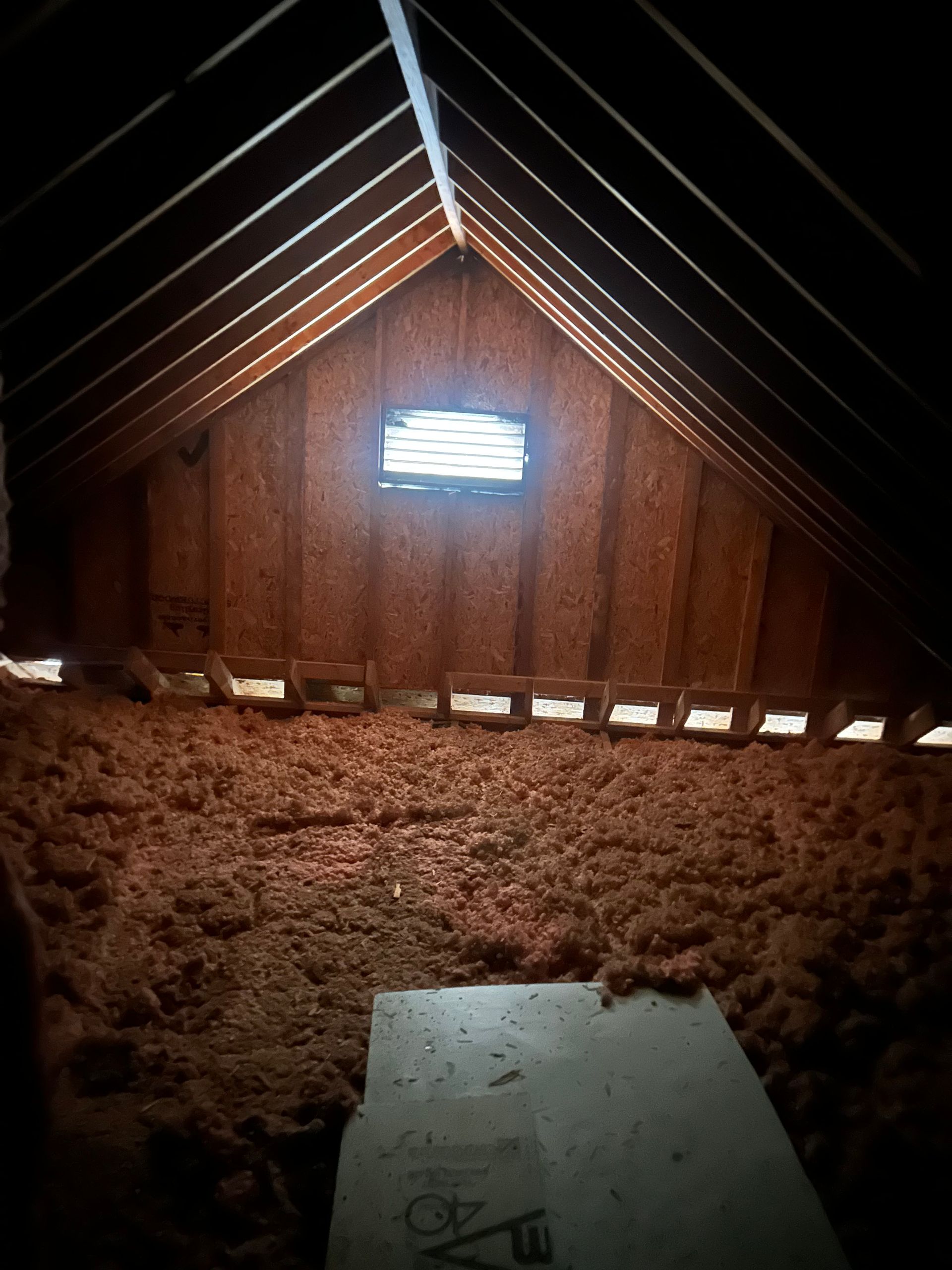 Attic space showing brown insulation covering the floor, a wooden framed ceiling, and a bright rectangular gable vent.