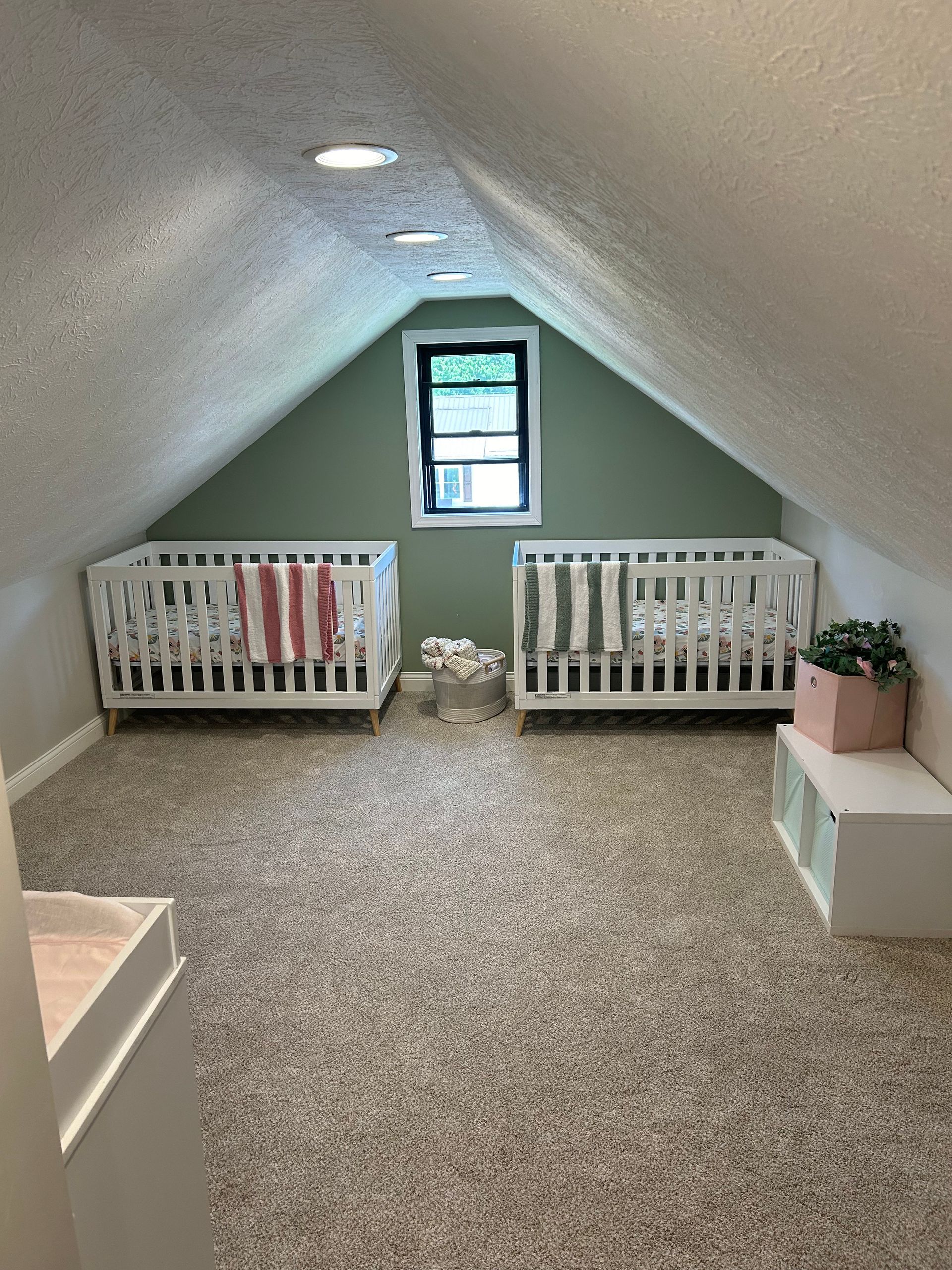 An attic nursery with two white cribs, green walls, a window, and a white storage unit on beige carpet.