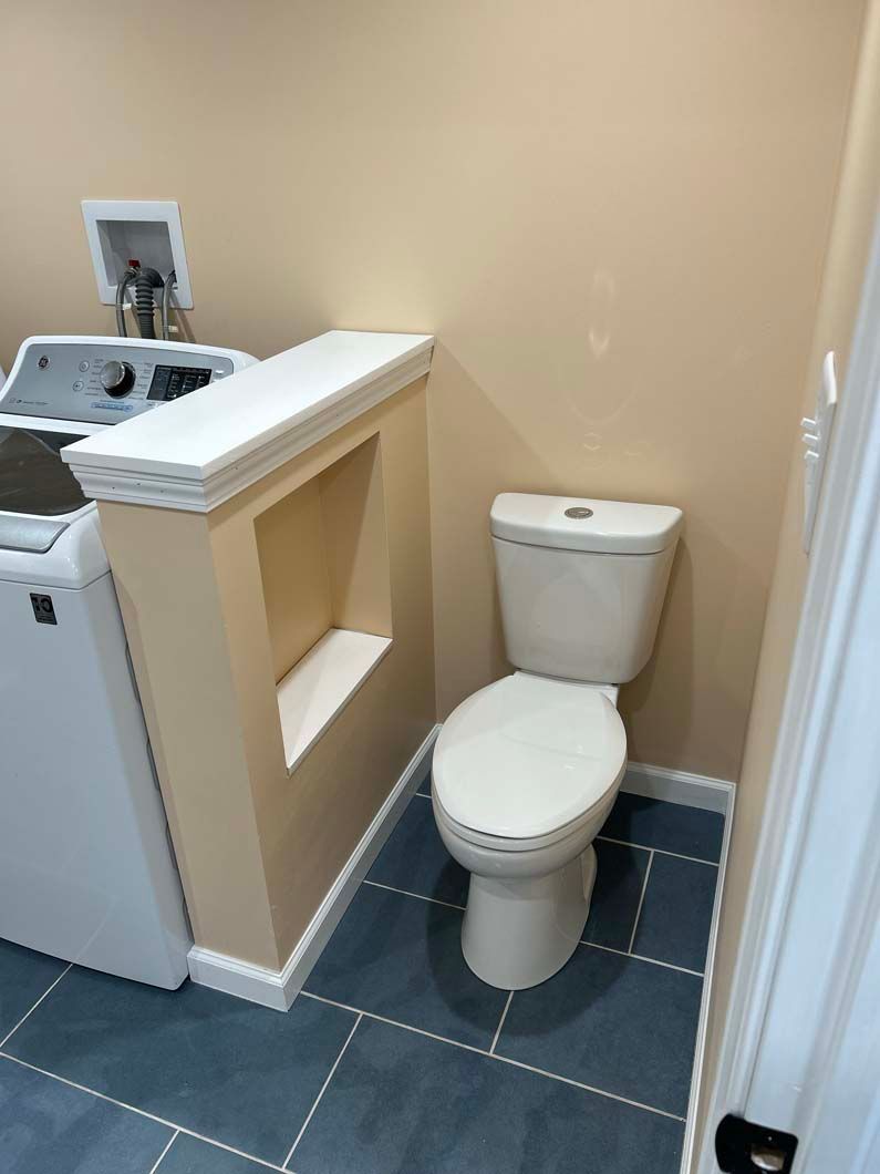 A beige toilet sits in a small room next to a half-wall partition featuring a built-in shelf and a laundry machine.
