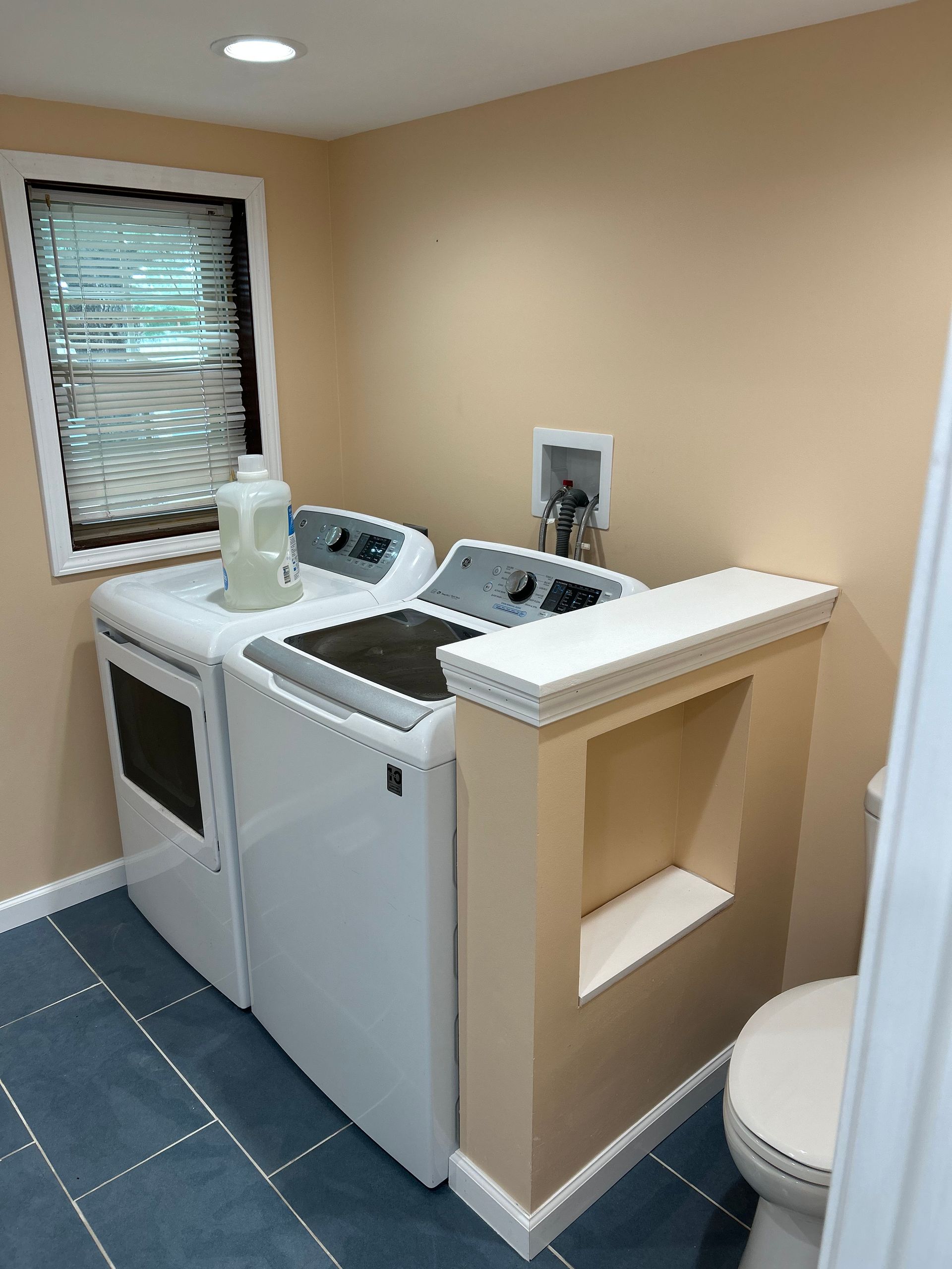 A laundry area with a white washer and dryer next to a tan partial wall with a recessed shelf and a toilet nearby.