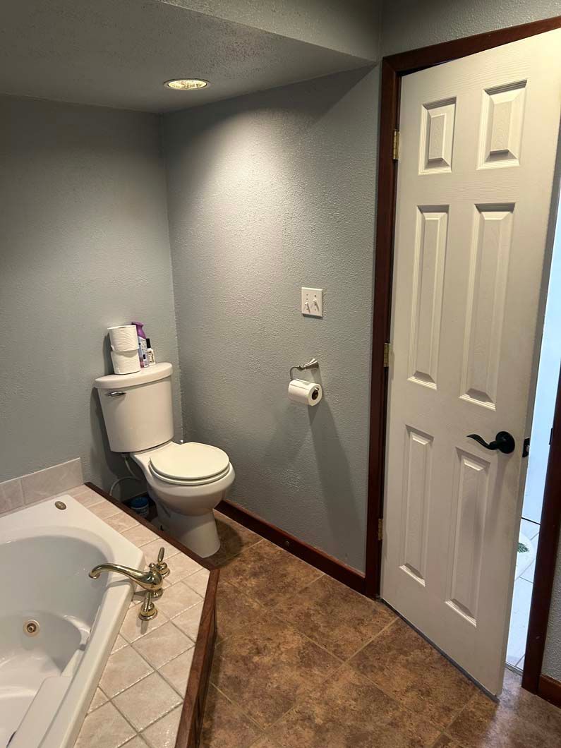 A bathroom showing a toilet next to a corner tub, with a white door and a light blue wall.