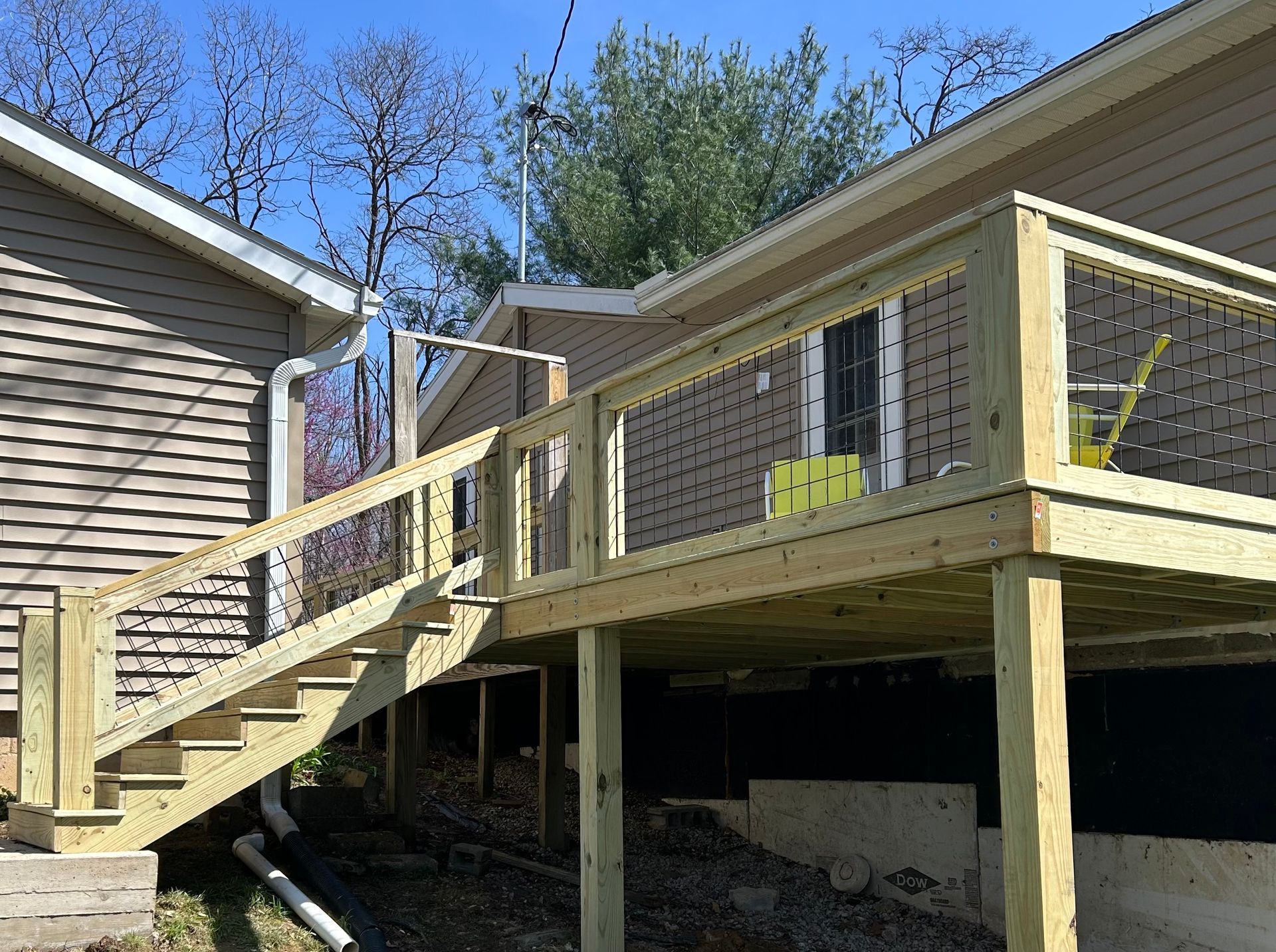 A newly built wooden deck with a staircase and cable railing stands next to a house with beige siding.