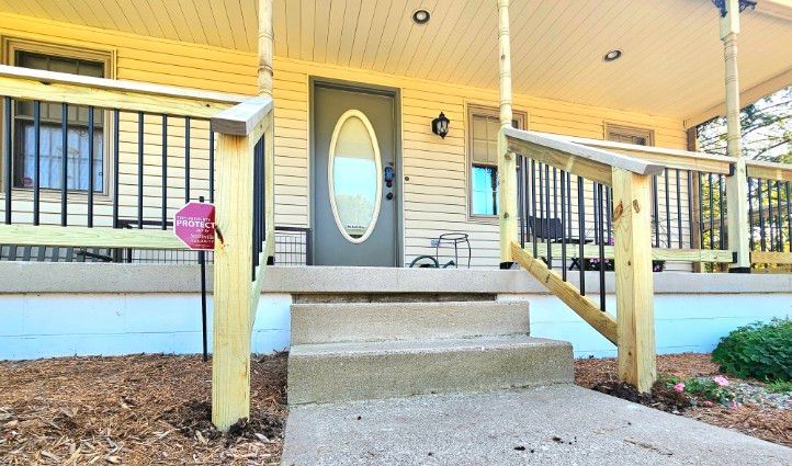 Concrete steps lead to the front porch of a yellow house with a grey oval-window door and wooden railings.