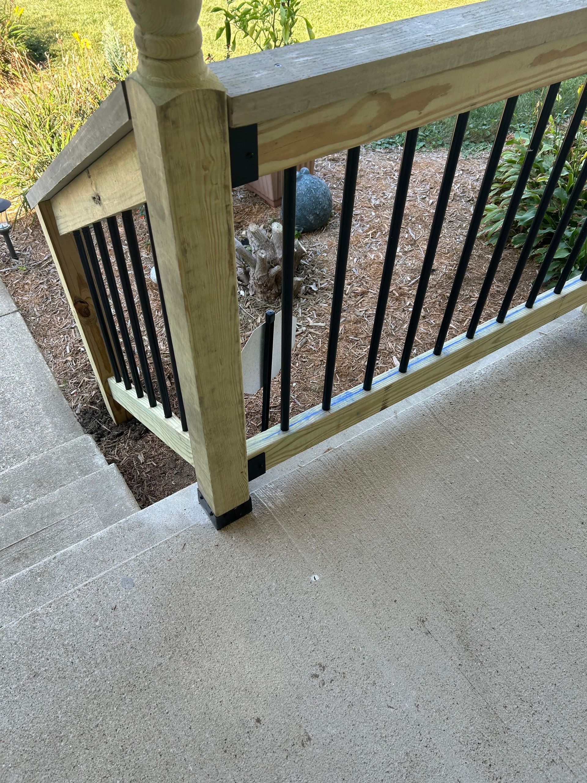 A wooden porch railing with black metal spindles, featuring a post corner mounted on a textured concrete step surface.