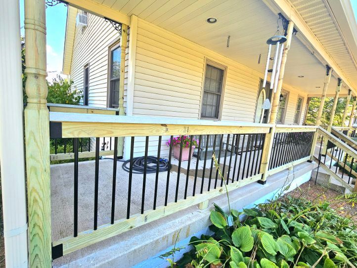 A yellow-sided house with a wooden front porch featuring black railings and a concrete floor, framed by green plants.
