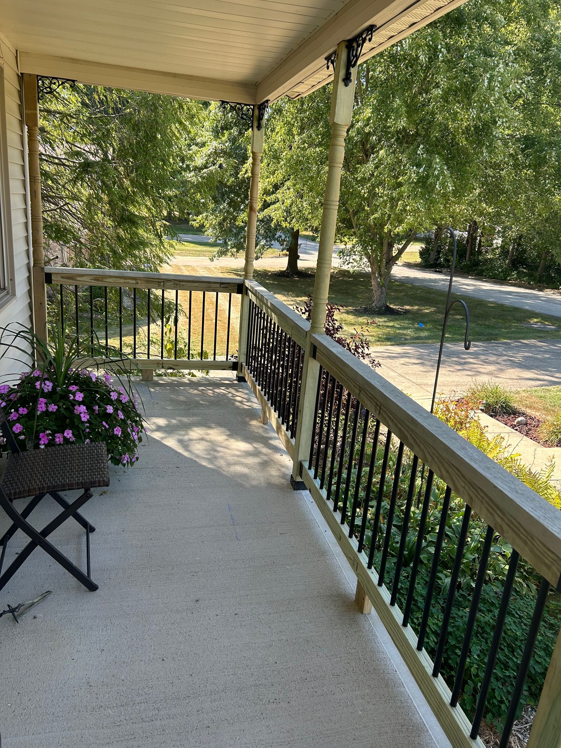 A wooden porch with black metal railings overlooks a sunlit garden with trees and greenery.